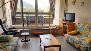 Ein Wohnzimmer mit einer Couch, einem Sessel, einem Tisch, einem Fernseher und Blick auf einen Balkon mit Gartenmöbeln.
