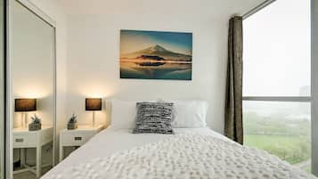 A hotel room with a large bed, a nightstand with lamps, a painting of a mountain, and a view of a park through the window.