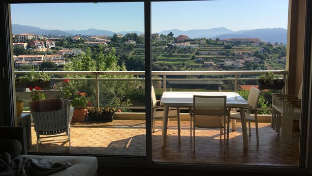 Un balcó amb taula i cadires, plantes en testos i vista a un paisatge ondulat ple d'edificis.