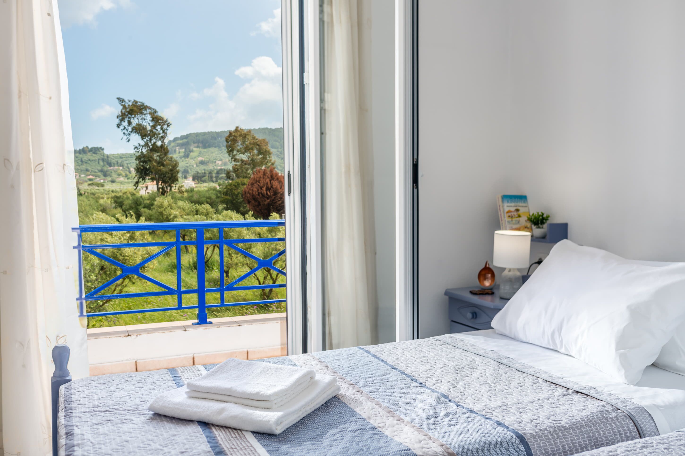 Une chambre à coucher comprenant un lit, une table de chevet, une lampe et une vue sur la verdure, accessible par un balcon doté de balustrades bleues.