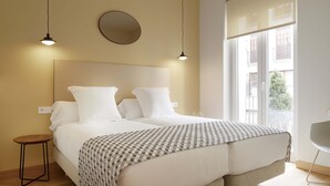 Ein Schlafzimmer mit einem großen Bett, zwei Nachttischen, einem runden Spiegel und einem Fenster mit Blick auf Gebäude.