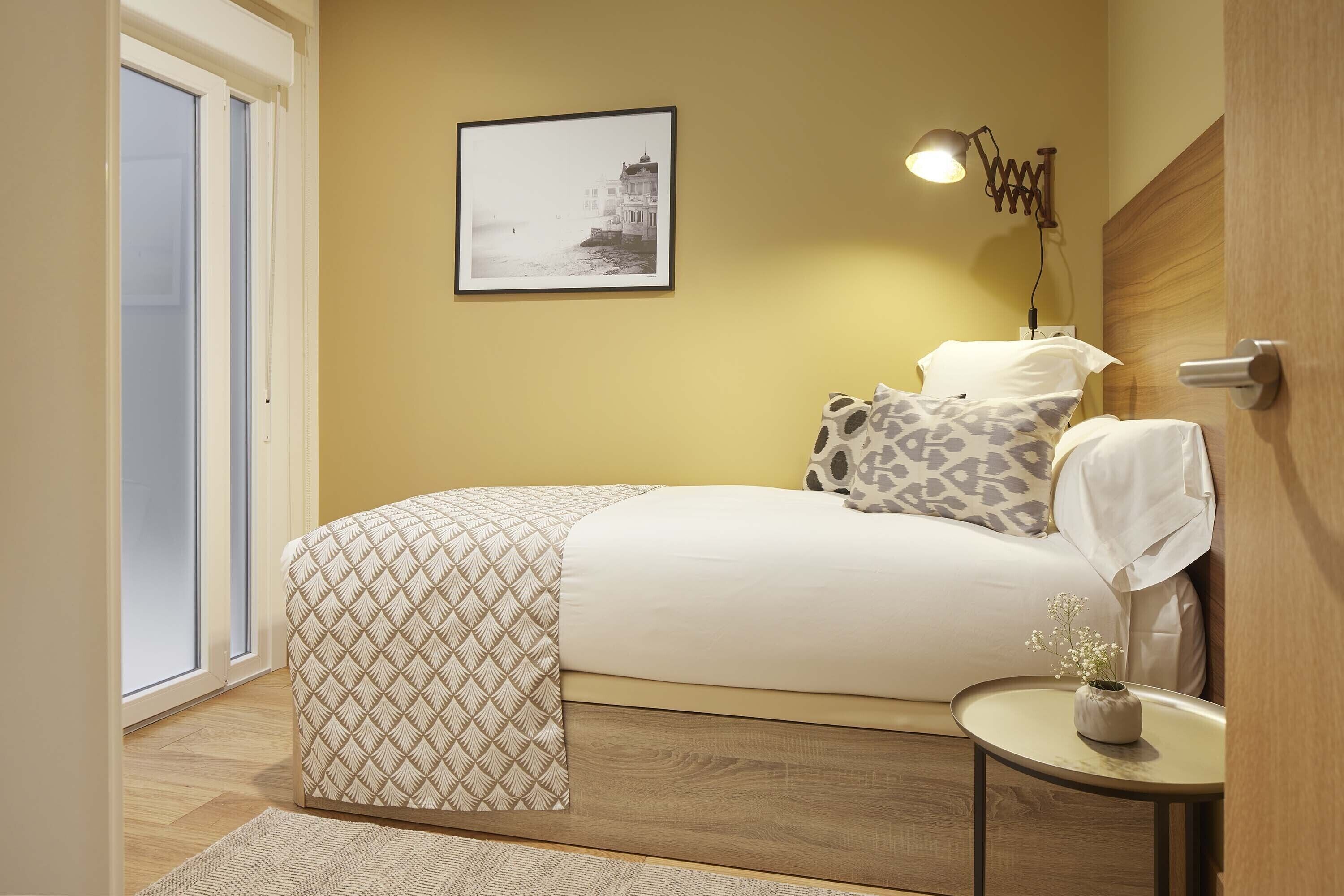 Une chambre à coucher avec un lit, une table de chevet avec une lampe, un tableau encadré au mur et une porte sur le côté gauche.