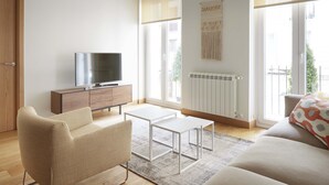 Ein modernes Wohnzimmer mit einer Couch, einem Couchtisch, einem Fernseher auf einer Holzkommode und großen Fenstern.