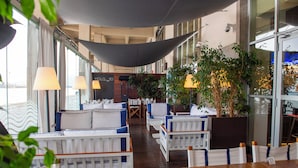 Un espacio interior con asientos de color blanco y cojines azules, ventanas amplias y plantas de interior.