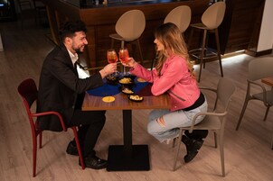 Una coppia seduta a un tavolo di legno con tovaglietta blu, che sorseggia delle bevande. Si notano sedie moderne e un'area bar sullo sfondo.