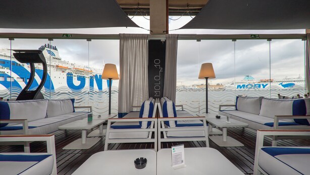Cubierta de crucero con zonas de estar, un ventanal con cortinas y vista del exterior del barco.