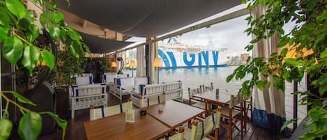 A restaurant deck with tables and chairs, overlooking a body of water and a cruise ship.