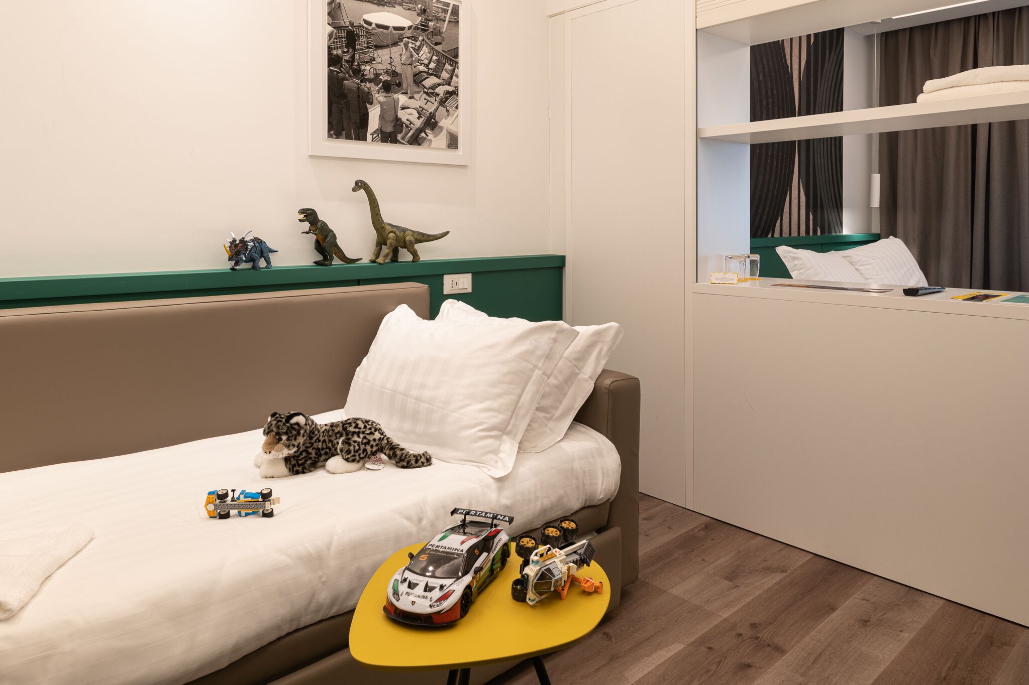 A hotel room with a bed, a small table with toys, a green shelf with dinosaur figurines, and a framed picture on the wall.