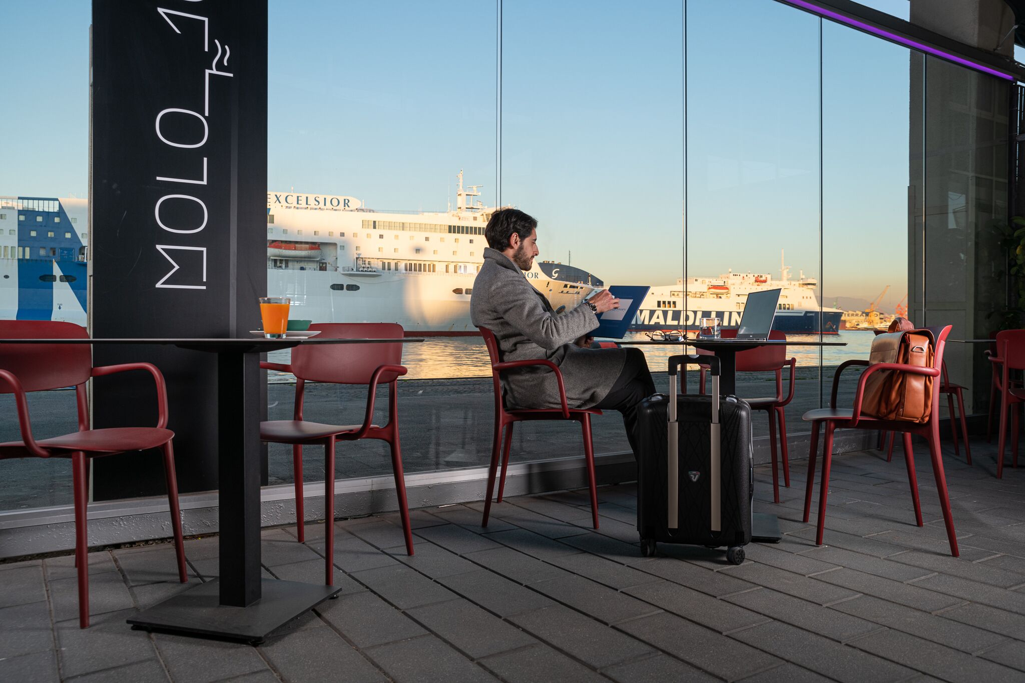 A man sitting at a table with a laptop, overlooking a body of water with ships in the background.