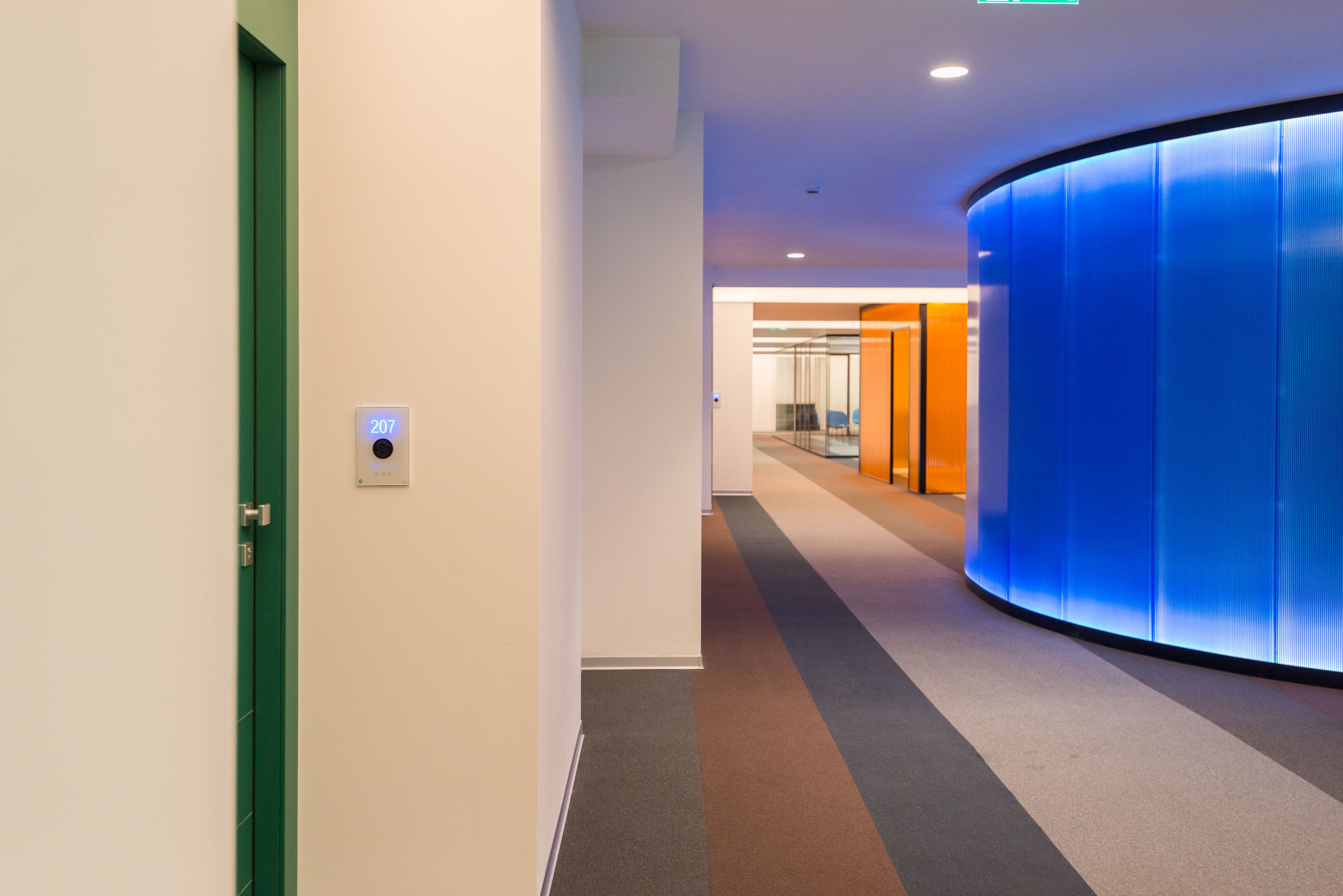 A modern hallway with a green door, a digital display showing the number 207, and a curved blue wall.