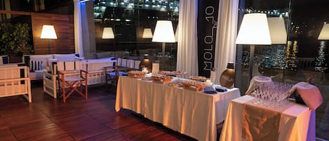 A dining area with a long table set for a meal, surrounded by chairs, and large windows with curtains.