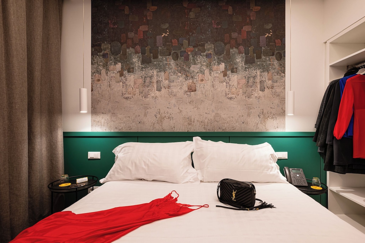Habitación de hotel con cama, almohadas blancas, una prenda roja, un bolso negro y una pared con textura.