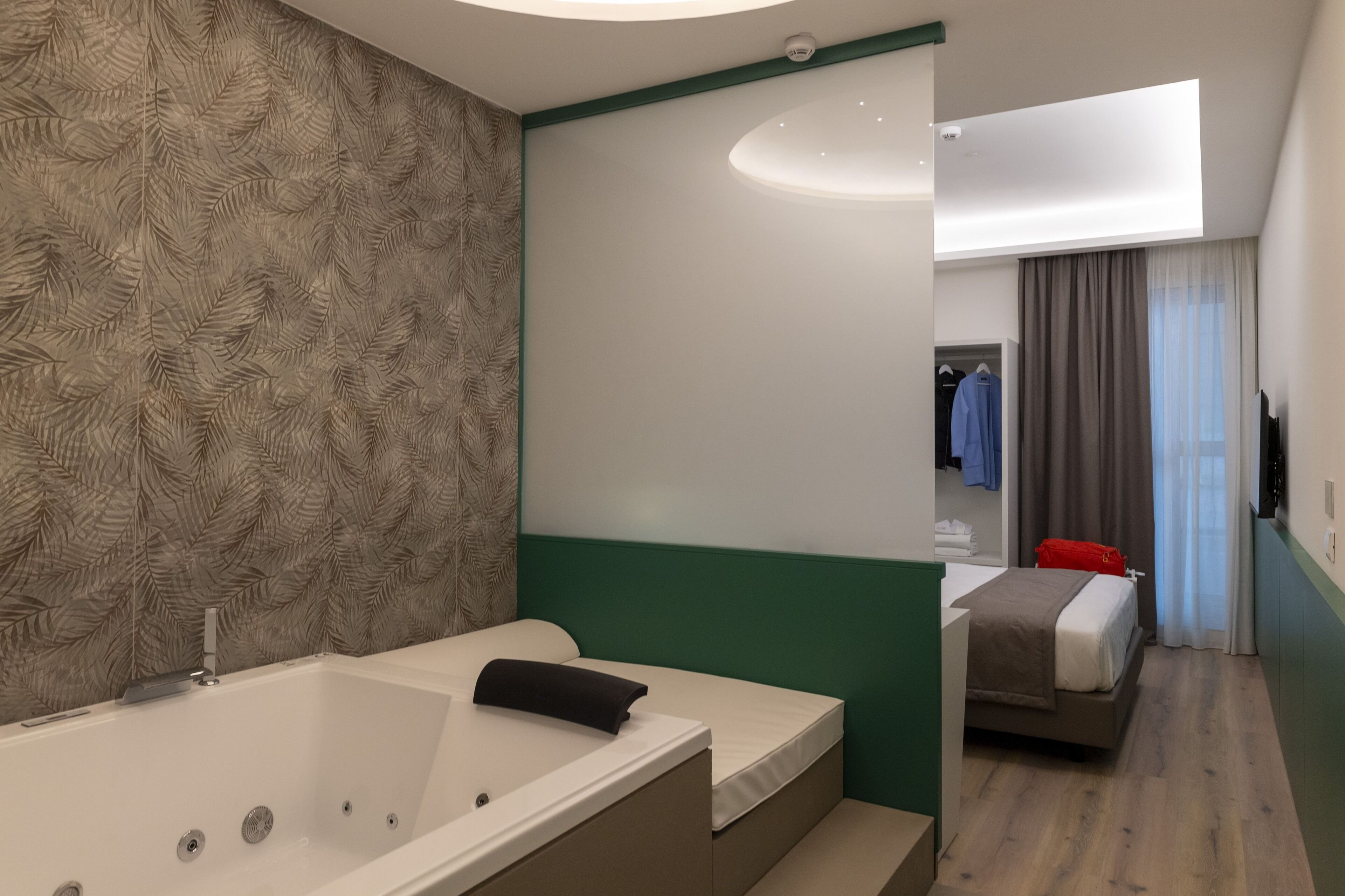 Una habitación de hotel moderna con una bañera grande, un dormitorio separado con cama, un espejo y vistas al pasillo.