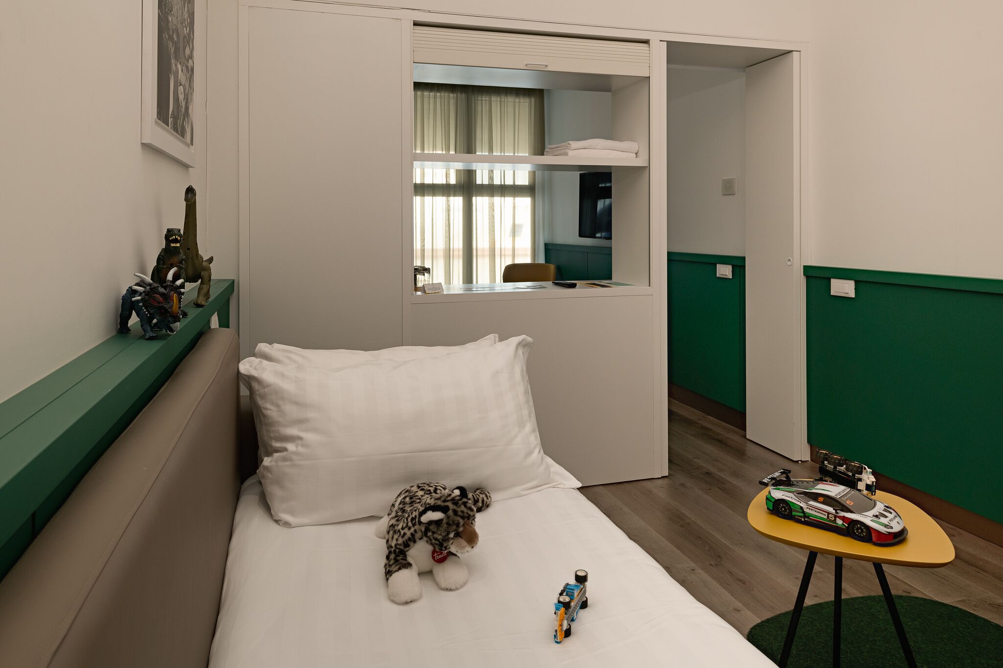 Una habitación pequeña y funcional con una cama, un banco verde, una mesita con juguetes y un espejo.