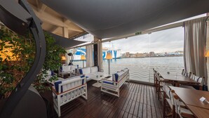 Una terraza con suelo de madera, asientos blancos y azules, y vista al agua y al paisaje urbano.