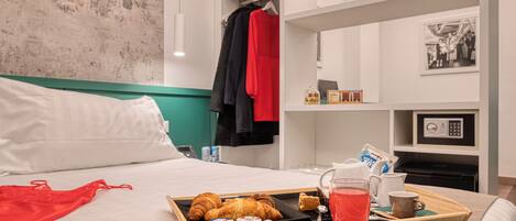 Cama bem arrumada com uma bandeja de itens de café da manhã, incluindo croissants, uma bebida e uma caneca, em um quarto moderno com uma parede de destaque verde e um casaco pendurado na porta.