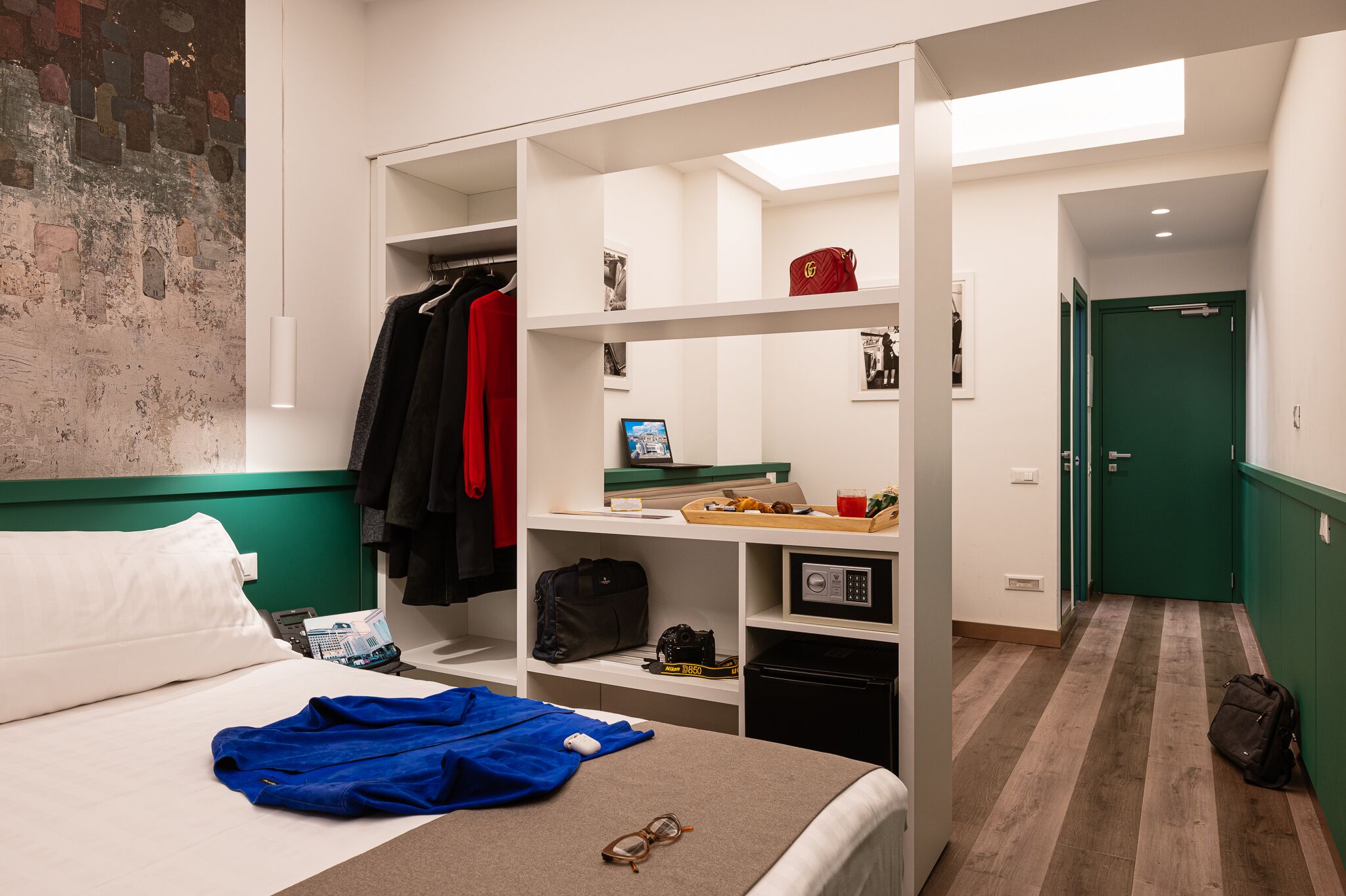 Quarto de hotel com cama, guarda-roupa, escrivaninha com computador e uma porta verde ao fundo.