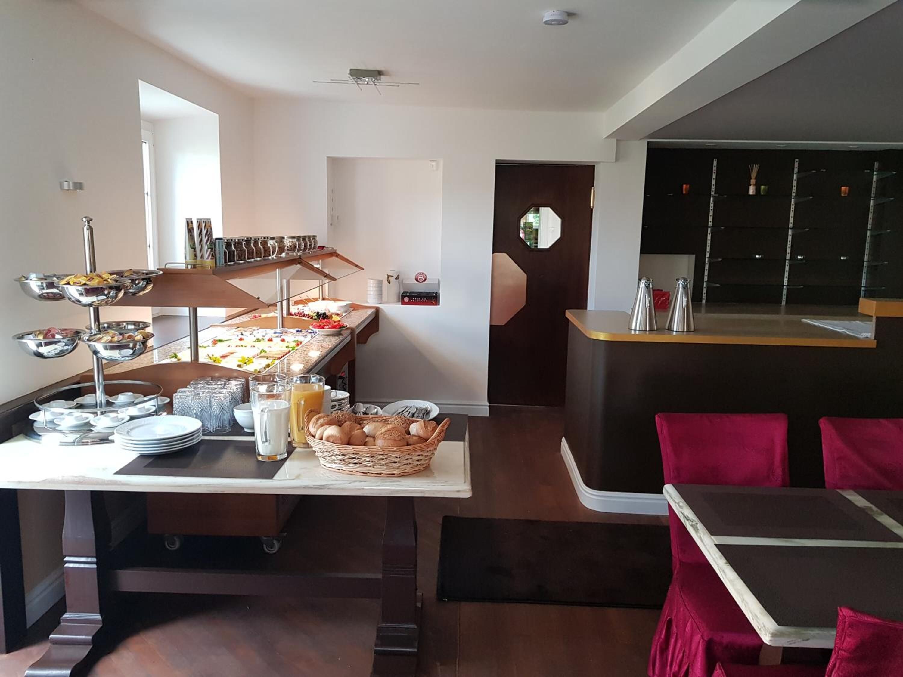 Ein reichhaltiges Frühstücksbuffet mit verschiedenen Speisen und Getränken, eine Kaffeestation und eine Sitzecke mit roten Kissen.