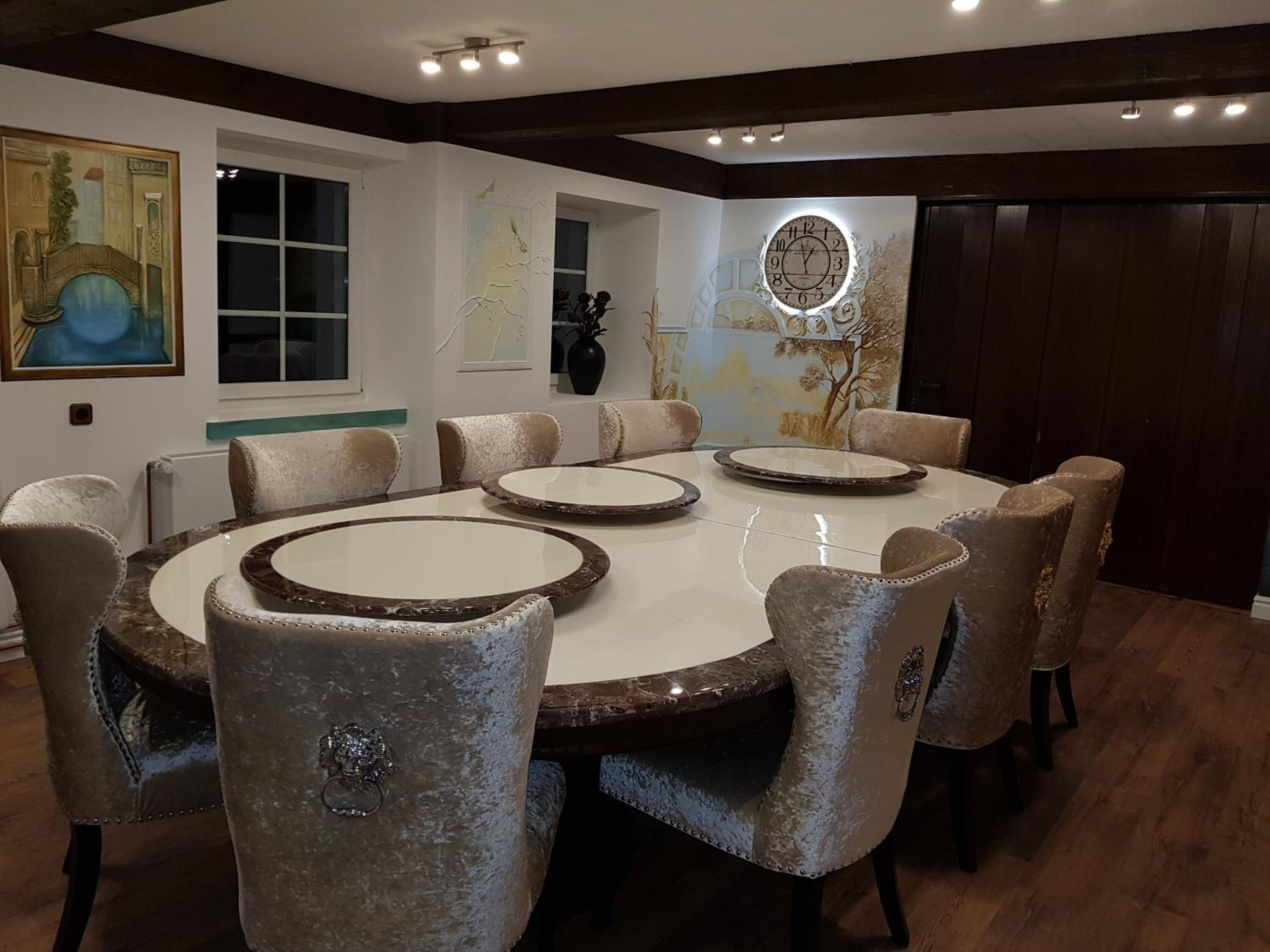 Un comedor con una mesa redonda y ocho sillas, un cuadro en la pared y un reloj.