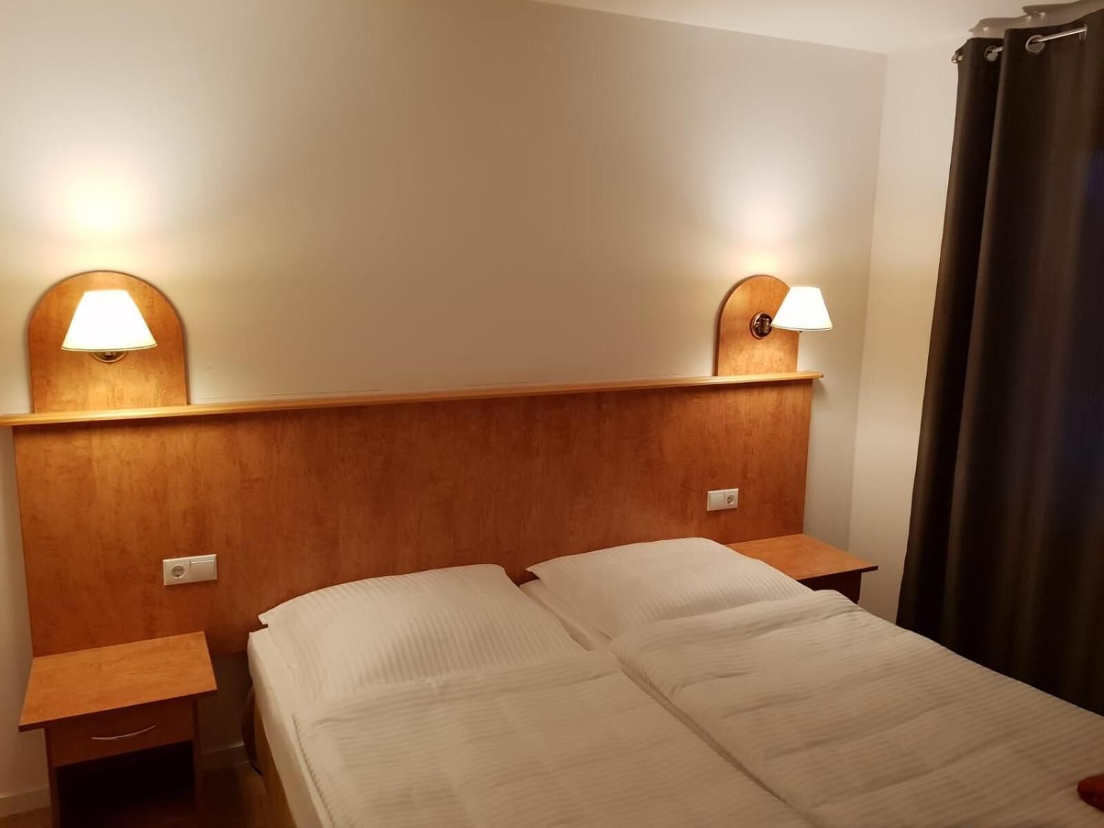 Habitación de hotel con cabecera de madera, dos lámparas de noche, cama con ropa blanca y cortina oscura en la ventana.