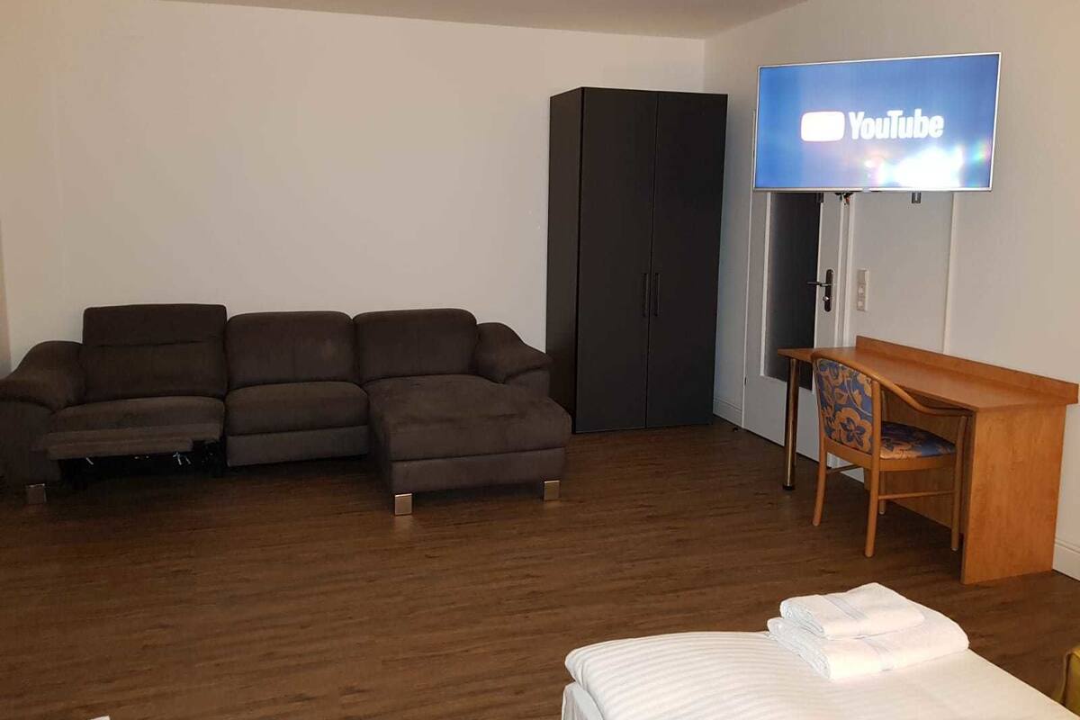Quarto de hotel moderno com sofá modular, escrivaninha de madeira, televisão de tela plana na parede e cama com roupa de cama branca e bem arrumada.