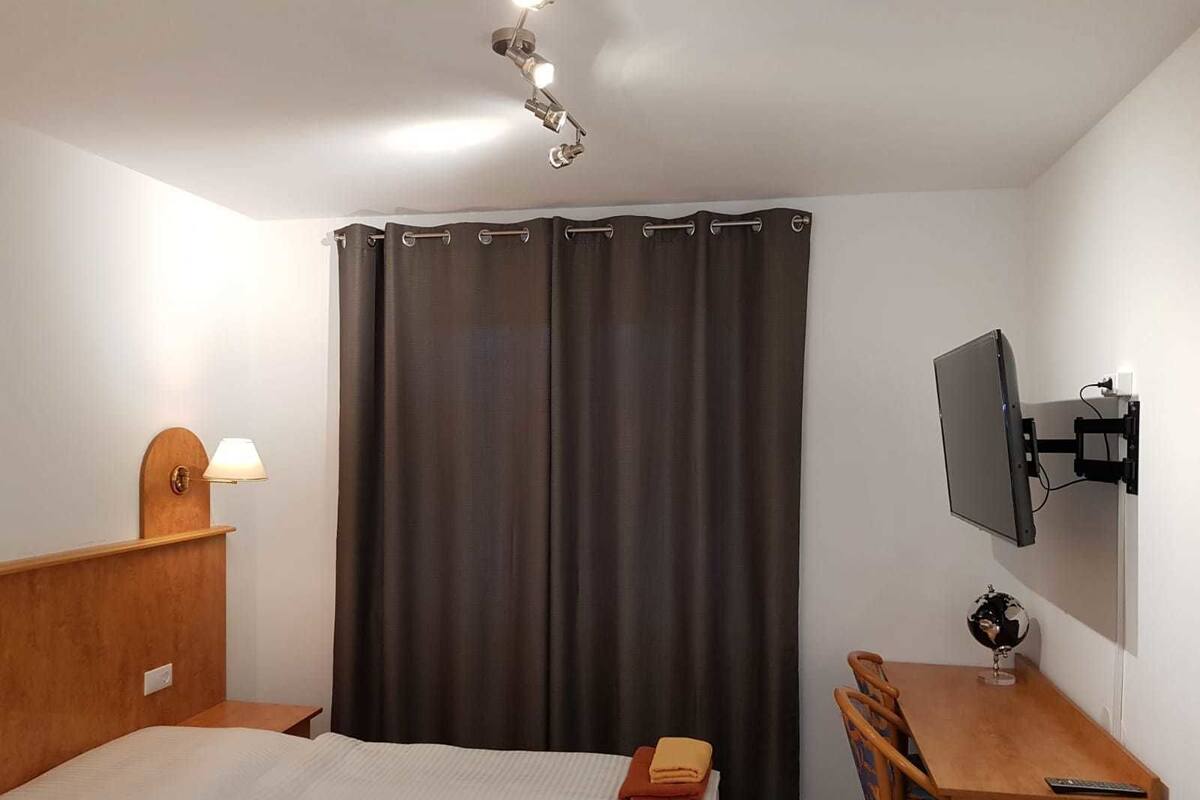 Quarto de hotel com cama, escrivaninha com cadeira, televisão de tela plana na parede e cortina.