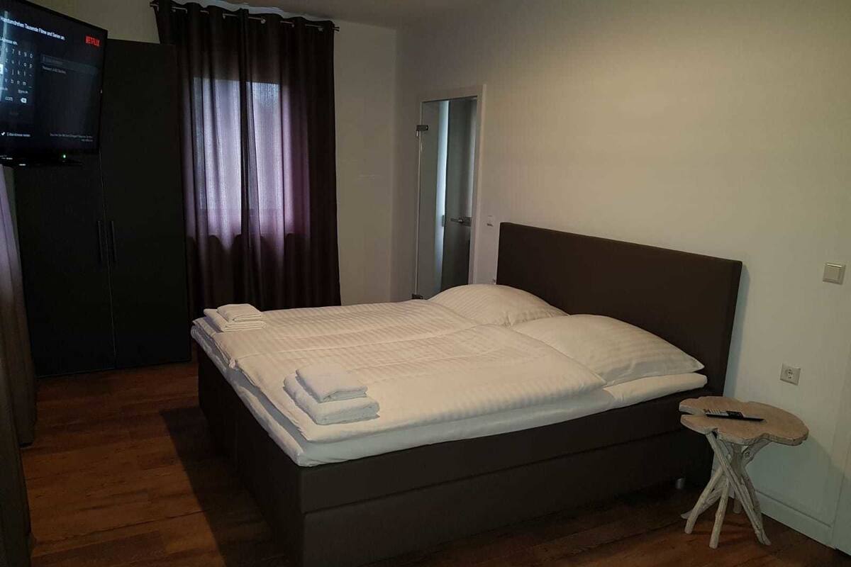Uma cama bem arrumada com roupa de cama branca, uma pequena mesa de cabeceira com um controle remoto e uma televisão fixada na parede.