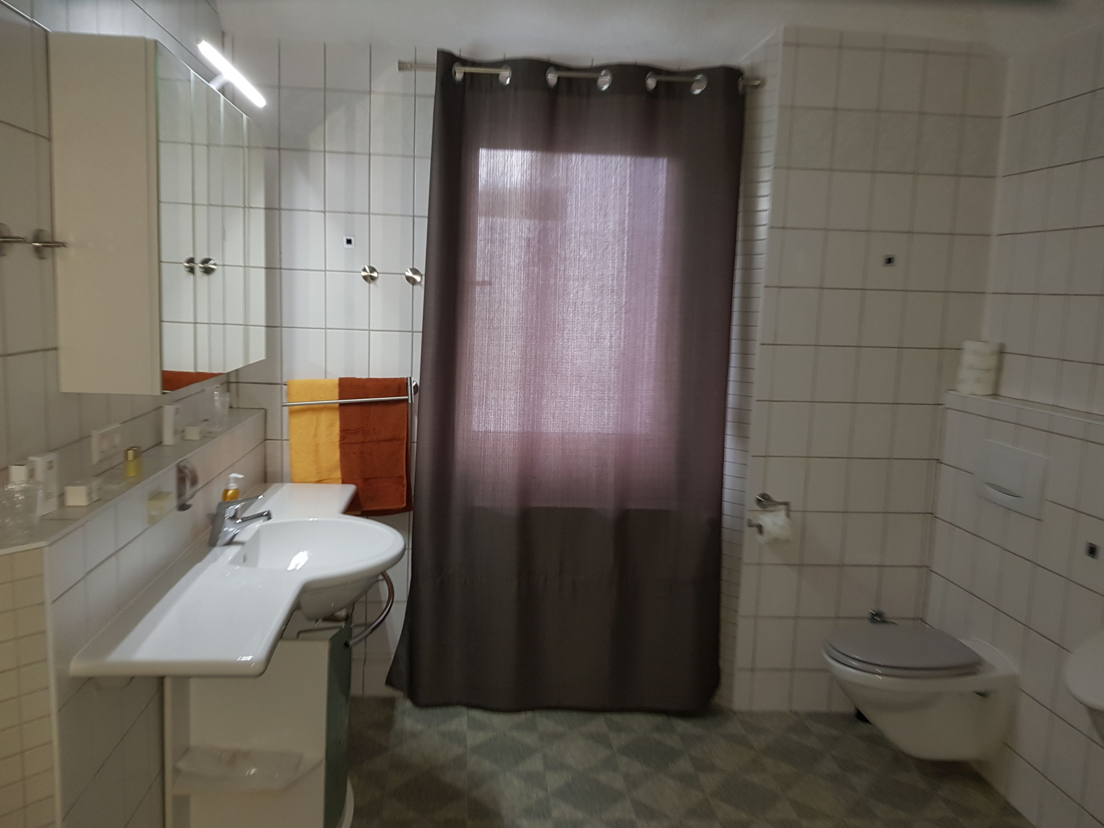 Een badkamer met een witte wastafel, spiegel en toilet. Er is een douchegordijn en een handdoekrek met handdoeken.