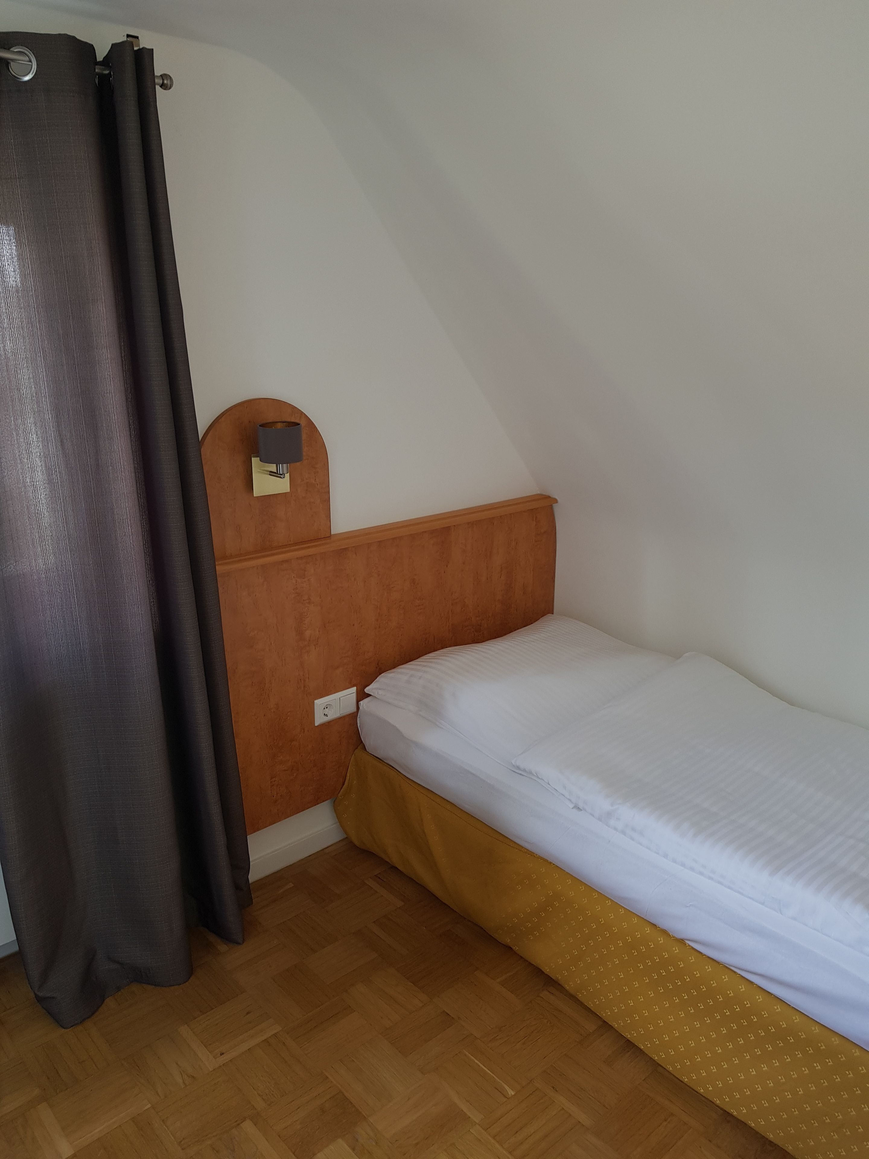 A small, single-bed room with a wooden headboard, a wall-mounted lamp, and a curtain.