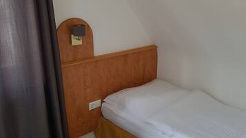 A small, single-bed room with a wooden headboard, a wall-mounted lamp, and a curtain.