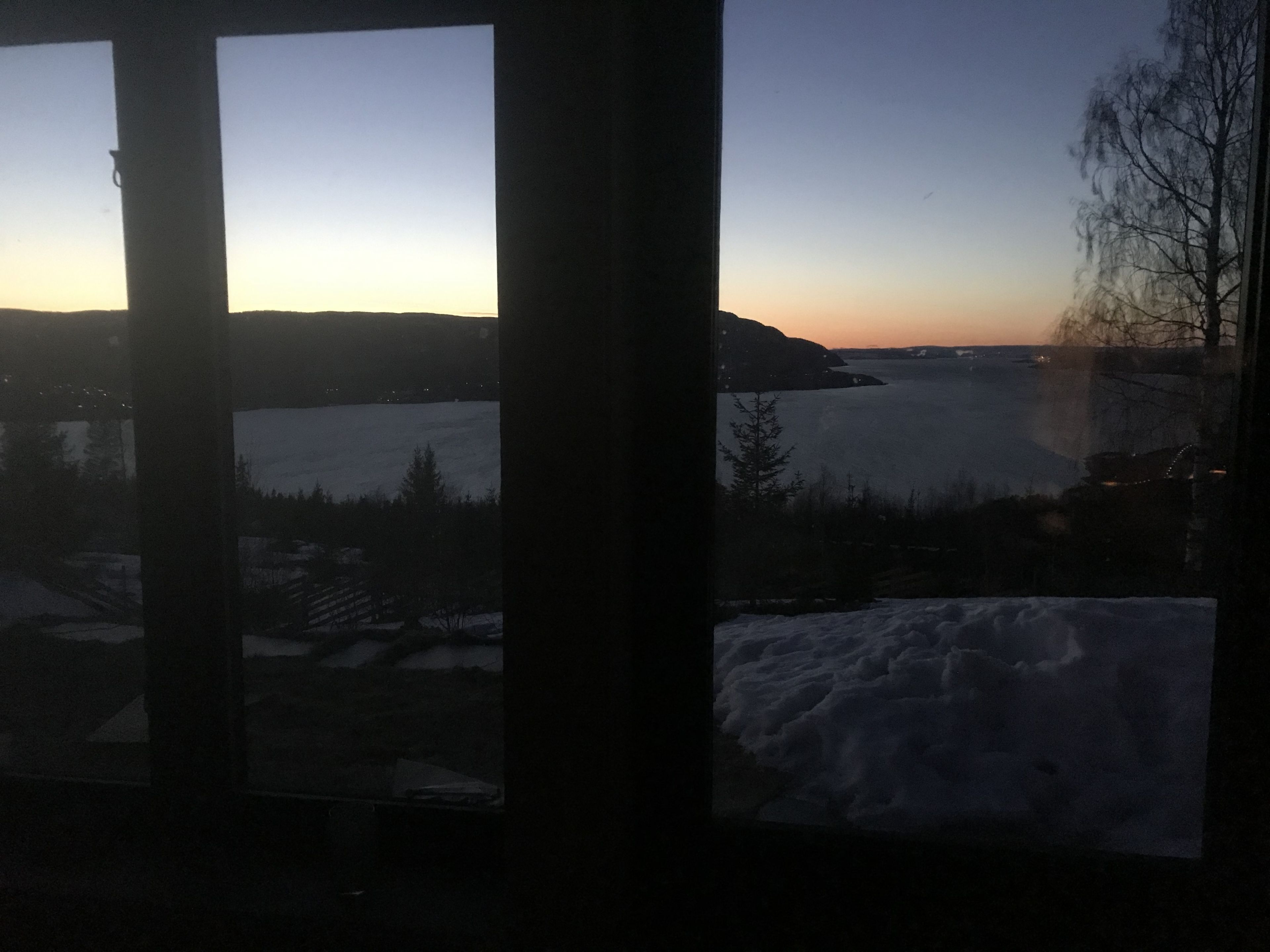 A view through a window with a snowy landscape, a body of water, and distant hills.