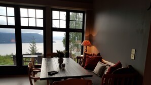 A room with a large window overlooking a lake, a wooden table, chairs, a sofa, and a lamp.