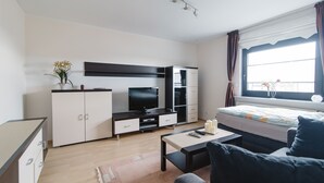 Une chambre moderne et bien éclairée, équipée d’un lit, d’une télévision, d’un bureau et d’une fenêtre avec des rideaux.