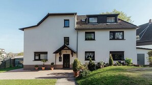 Une maison à deux étages, avec une façade blanche, un toit noir et un petit porche à l’avant, agrémenté de plantes en pot.