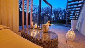 A cozy outdoor seating area with a wicker table, candles, and a champagne bottle. - La Douce Escapade (Sizun)