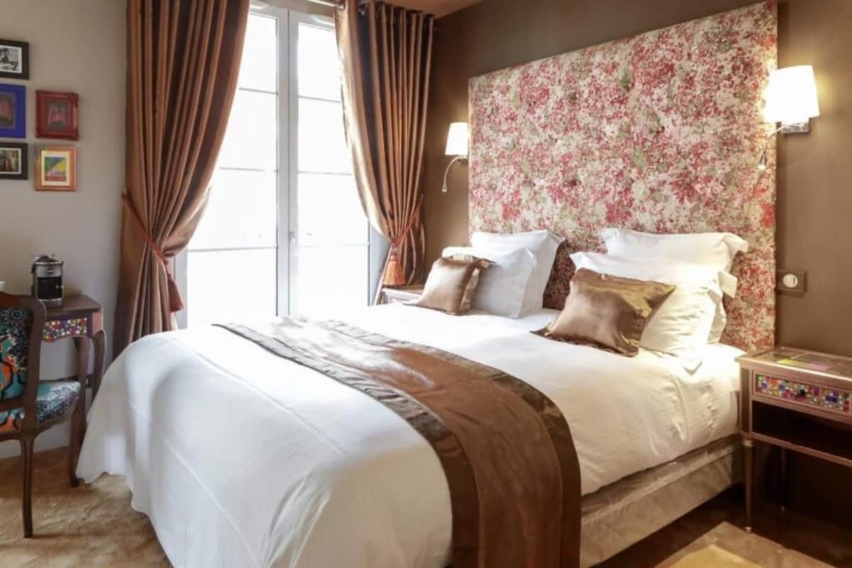 A neatly made bed with a floral headboard, a bedside table with a lamp, and a chair with a colorful cushion.