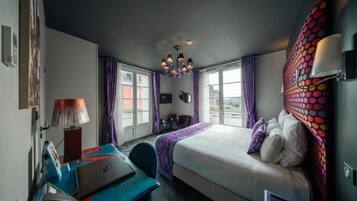 A hotel room with a bed, desk, chair, and a view of the city through large windows.