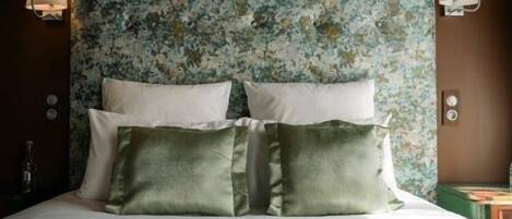 Una cama con sábanas blancas y almohadas verdes, cabecera con estampado floral y lámparas de pared a cada lado.