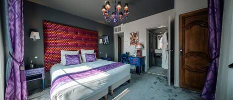 Un dormitorio con una cama grande, una araña, un cuadro de pared colorido y una puerta de madera.