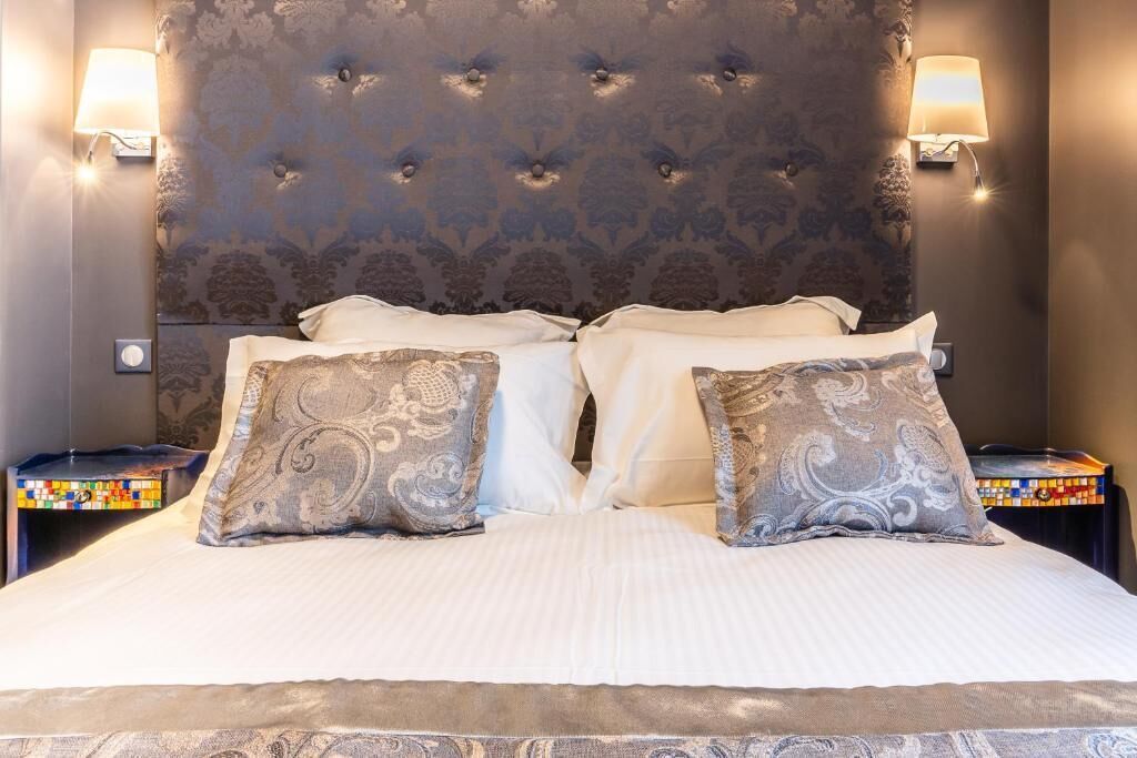 Una cama bien tendida con almohadas estampadas, un cabecero capitonado y lámparas fijadas a la pared a cada lado.