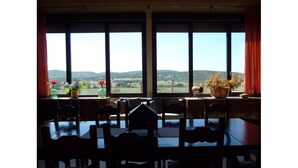 A dining area with a table and chairs, large windows with a view of hills, and potted plants on the windowsill. - Yeguada farm El Corral Delconde (Fernán Caballero)