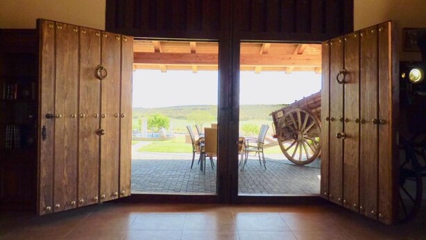 A wooden door with a view of a patio area, a table, and a wagon wheel. - Yeguada farm El Corral Delconde (Fernán Caballero)