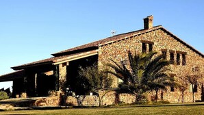 A stone building with a tiled roof, a covered porch, and a palm tree in front. - Yeguada farm El Corral Delconde (Fernán Caballero)