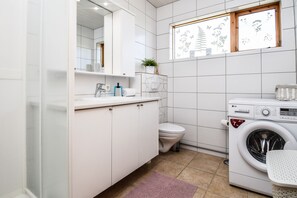 A modern bathroom with white tiles, a toilet, a washing machine, and a window with a plant on the windowsill. - House V10 (Bláskógabyggd)