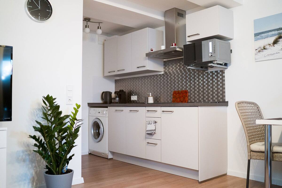 A modern kitchen with white cabinets, a built-in washing machine, and a dining area with a wicker chair.