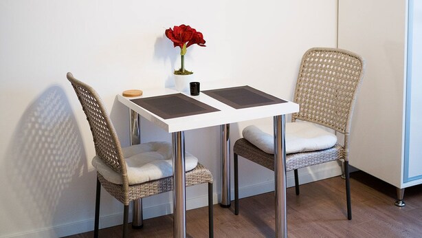 A dining area with a white table, two wicker chairs, a red flower vase, and three beach landscape paintings on the wall.