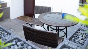 An outdoor seating area with wicker furniture and a glass table, surrounded by a stone pathway.