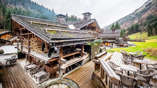 Eine Holzhütte mit einer Terrasse, Sitzgelegenheiten im Freien und einem Bergpanorama.