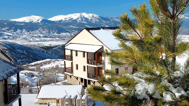 A multi-story building with balconies, surrounded by snow-covered mountains and pine trees. - Résidence Odalys Mille Soleils (Font-Romeu-Odeillo-Via)