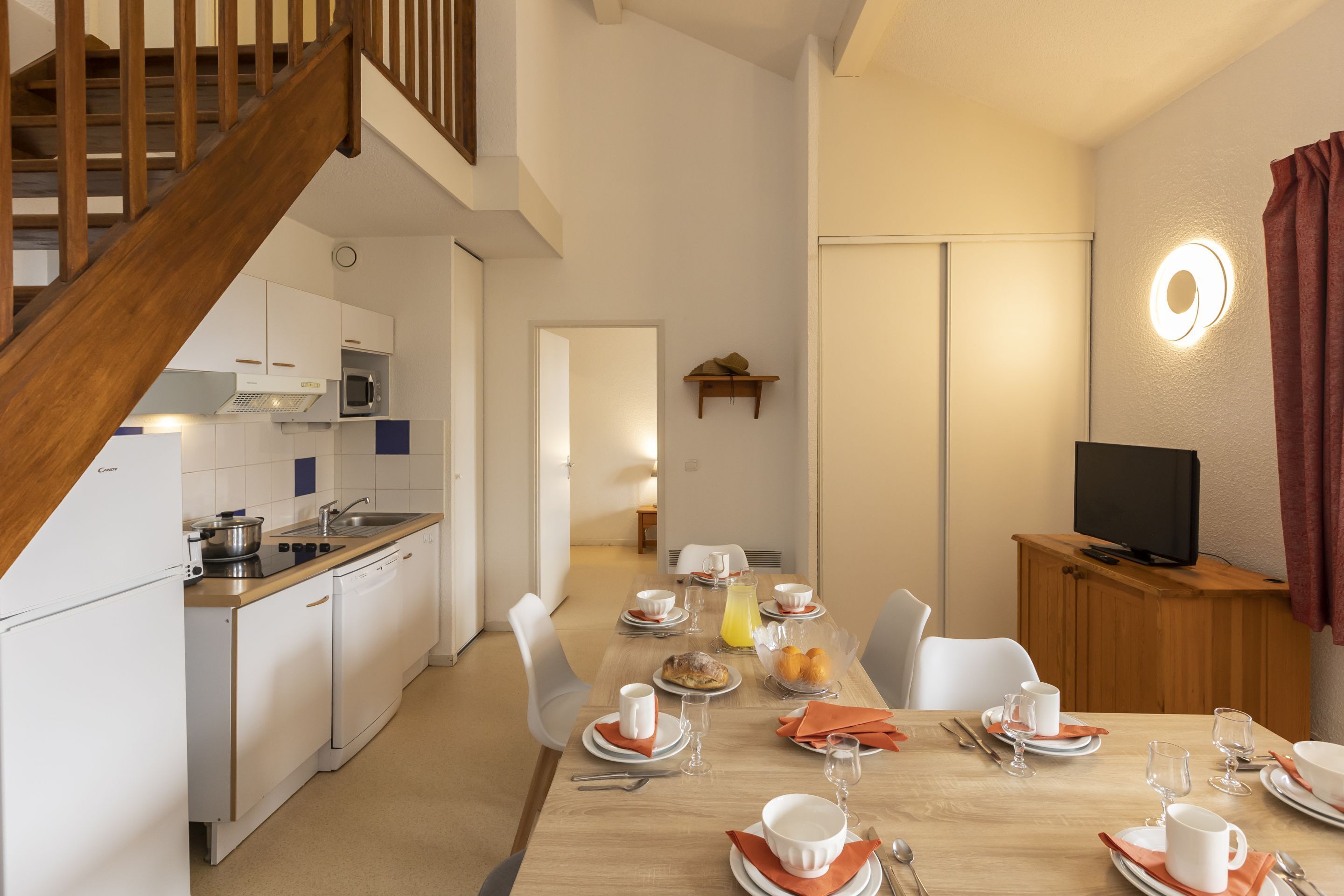 Une cuisine moderne avec des meubles blancs, un escalier en bois et un coin repas préparé pour le petit-déjeuner, avec des assiettes, des tasses et un panier de fruits.
